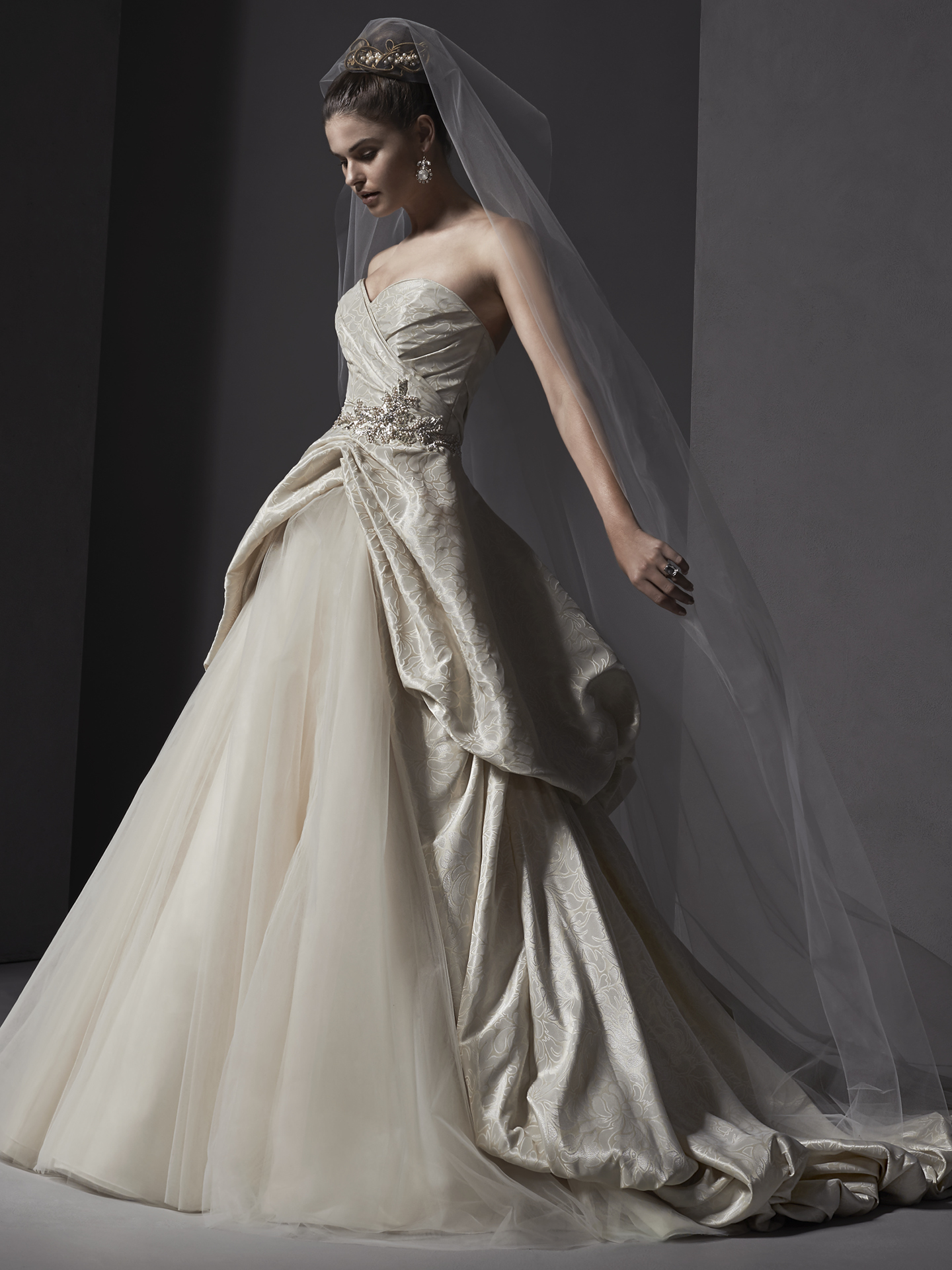 Brocade drapes the bodice of this ball gown before cascading into a voluminous cut-away skirt revealing tulle, accented with Swarovski crystal appliqué and sweetheart neckline. Finished with covered buttons over zipper and inner corset closure.